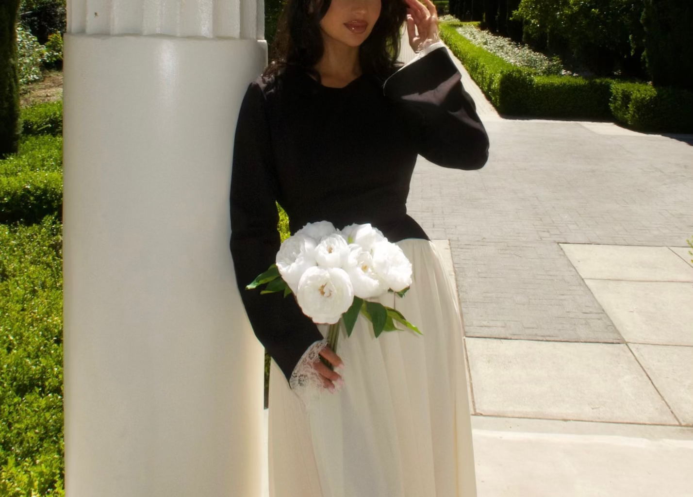 Woman in a modest black and white dress holding flowers, standing next to a column with trees in the background